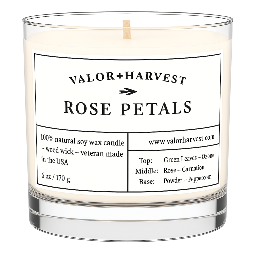 A clear glass soy wax candle with a label that reads 'Rose Petals'. It is on a wooden surface with bokeh lights in the background.