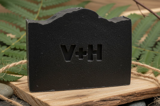 Black Forge activated charcoal soap bar by Valor+Harvest on a wooden slab with green ferns.